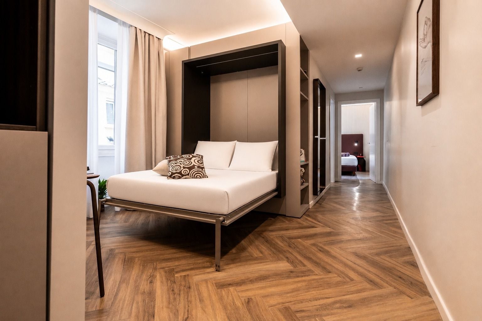 A modern hotel room with a bed, a wardrobe, and a hallway leading to another room.
