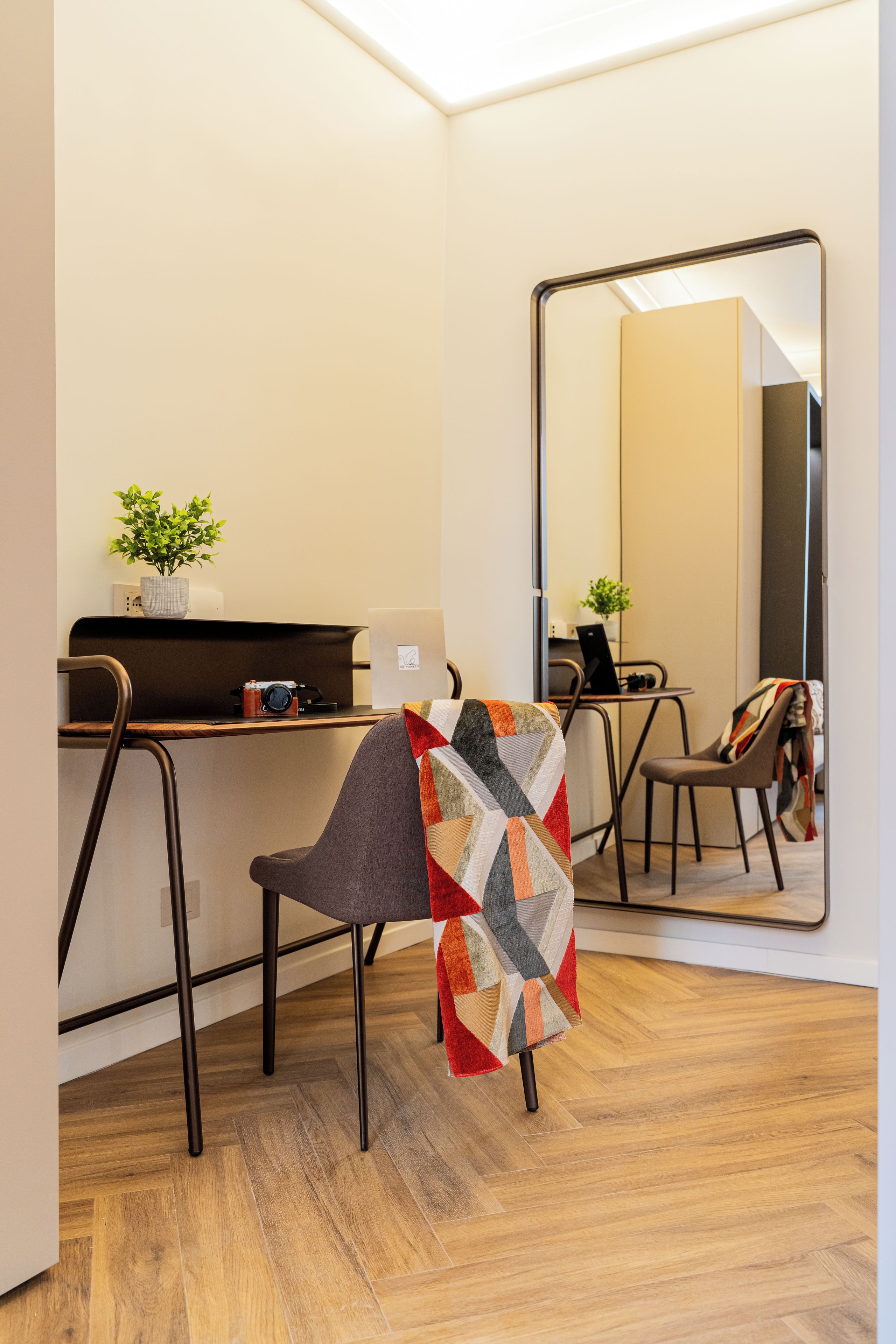 Quarto moderno com piso de madeira, uma escrivaninha com computador, uma cadeira com manta estampada e um espelho grande.