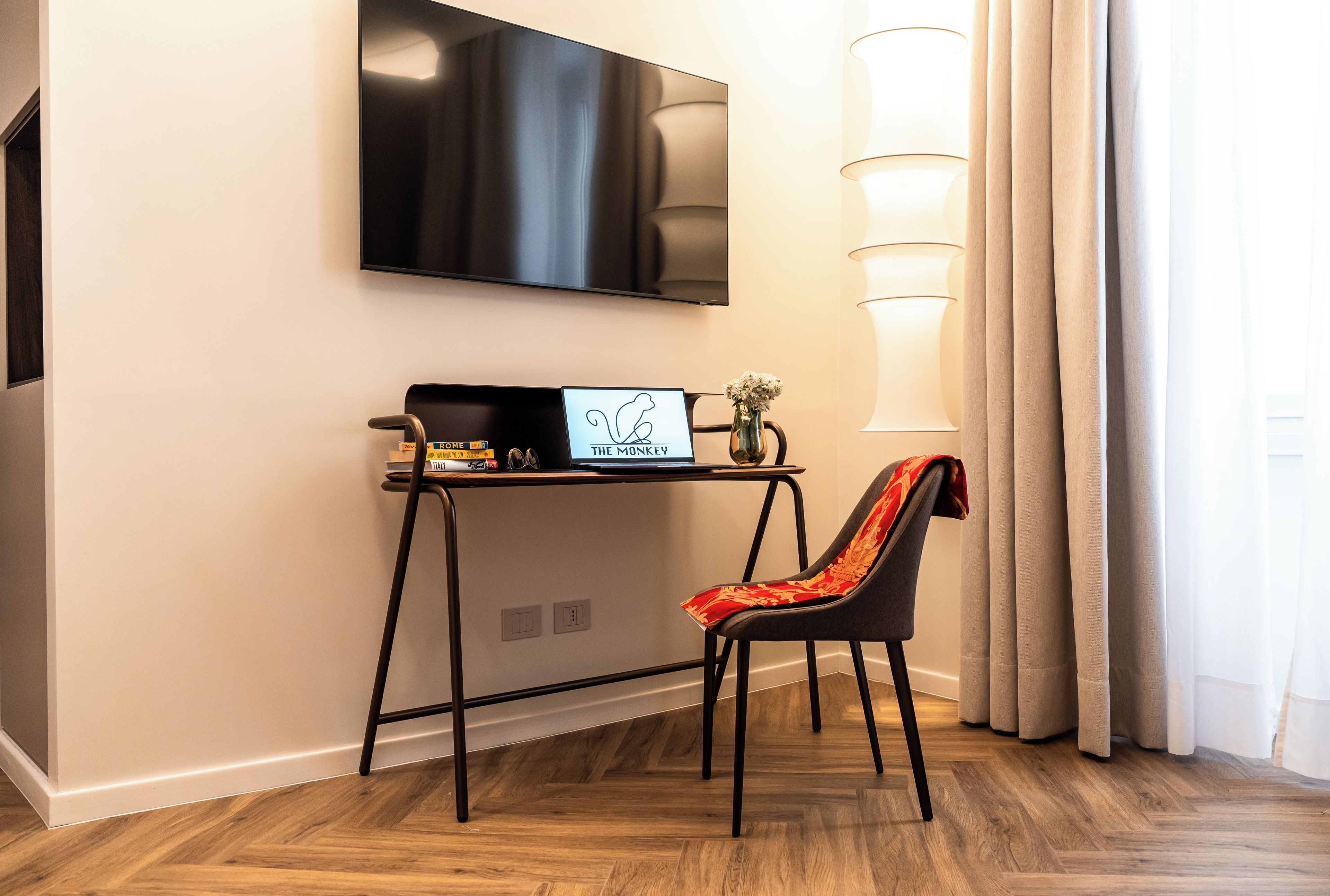 Quarto moderno com televisão de tela plana na parede, uma pequena mesa de trabalho com um laptop e um vaso de flores, uma cadeira com almofada vermelha, e uma janela grande com cortinas.