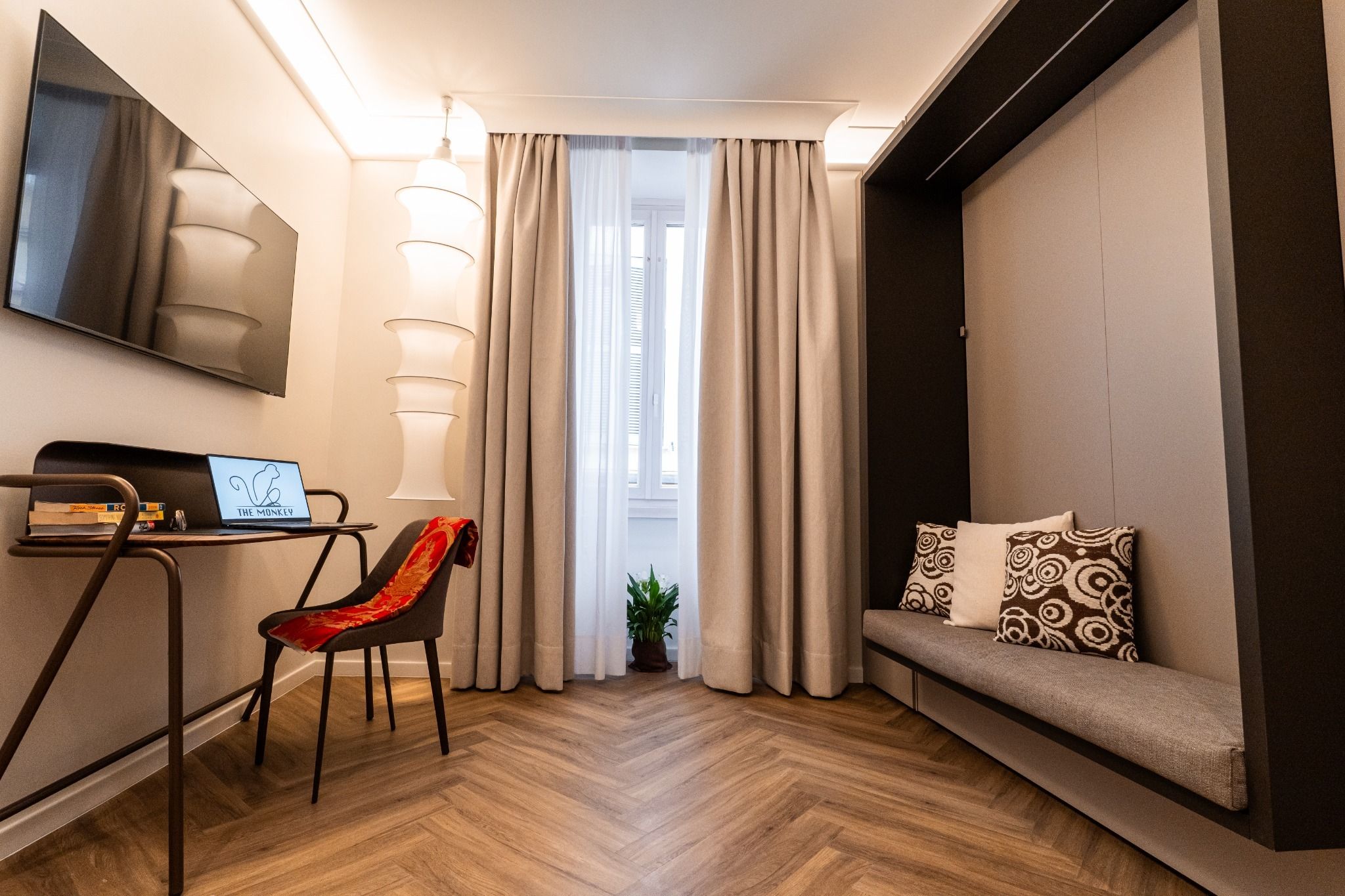 Quarto de hotel moderno com televisão de tela plana, escrivaninha com laptop, cadeira vermelha, sofá com almofadas estampadas e planta em vaso.