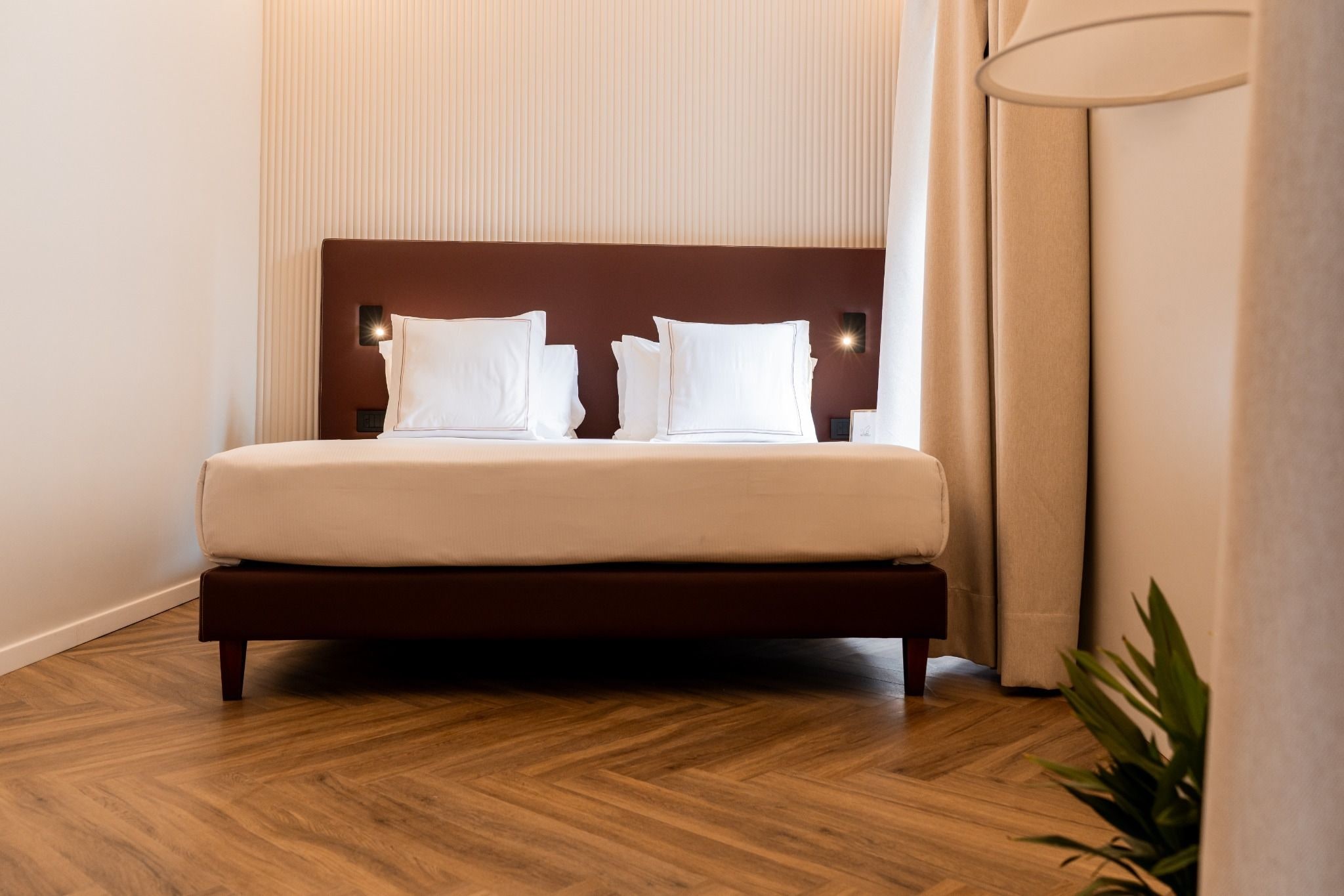 Un lit bien fait, avec une tête de lit en bois, une lampe de chevet et une plante en pot, dans une chambre d’hôtel.