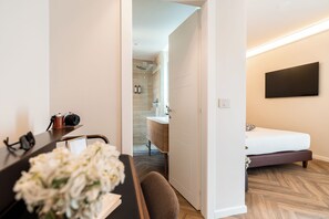 Une chambre d’hôtel avec un lit, un bureau équipé d’une caméra, une chaise, un téléviseur fixé au mur et une salle de bain visible à travers une porte ouverte.