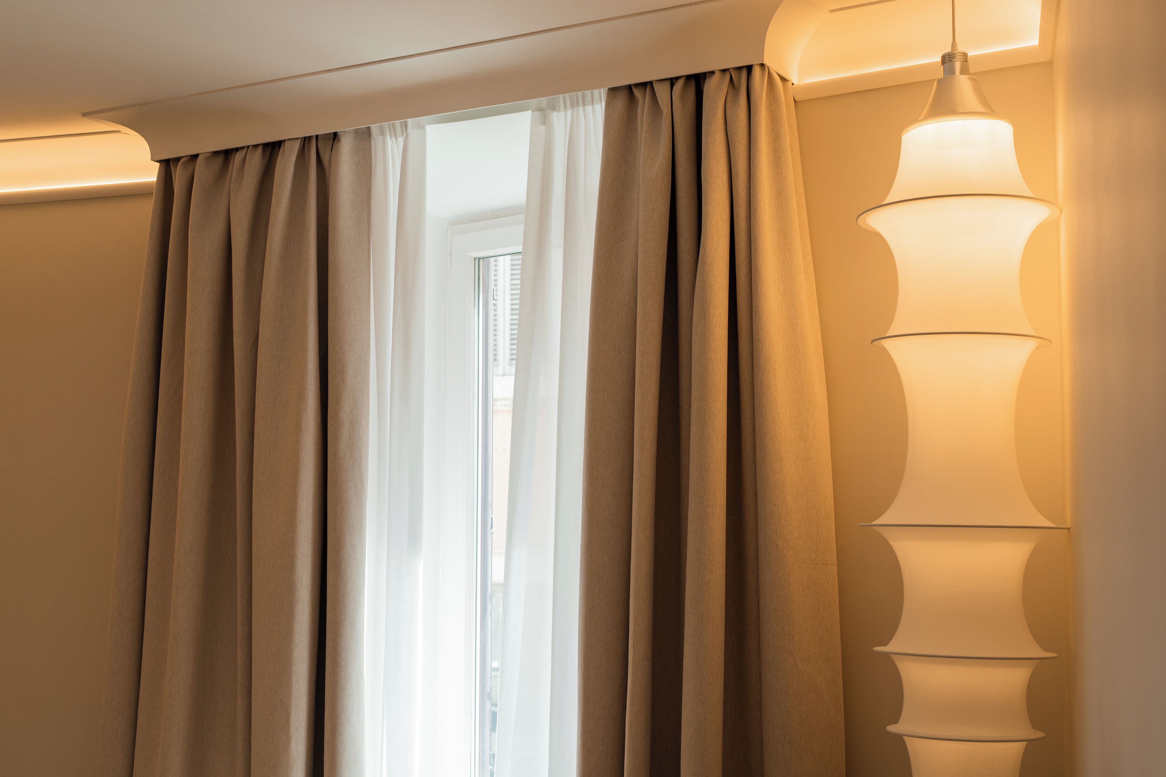 Quarto com janela coberta por cortinas bege, luminária fixada na parede e moldura de porta visível.