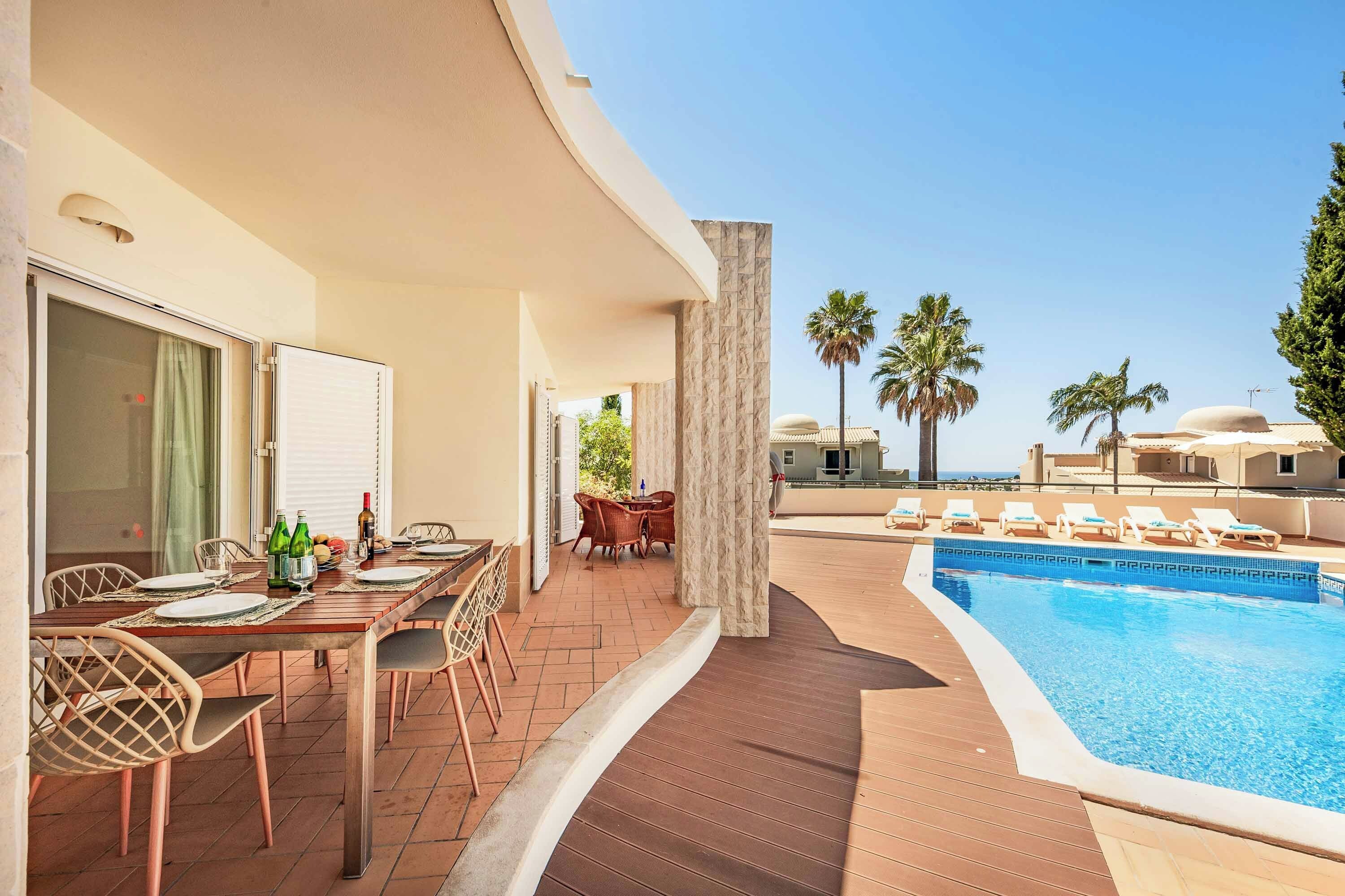 A poolside area with a dining table set for two, a pool with lounge chairs, and a view of palm trees and buildings in the distance.