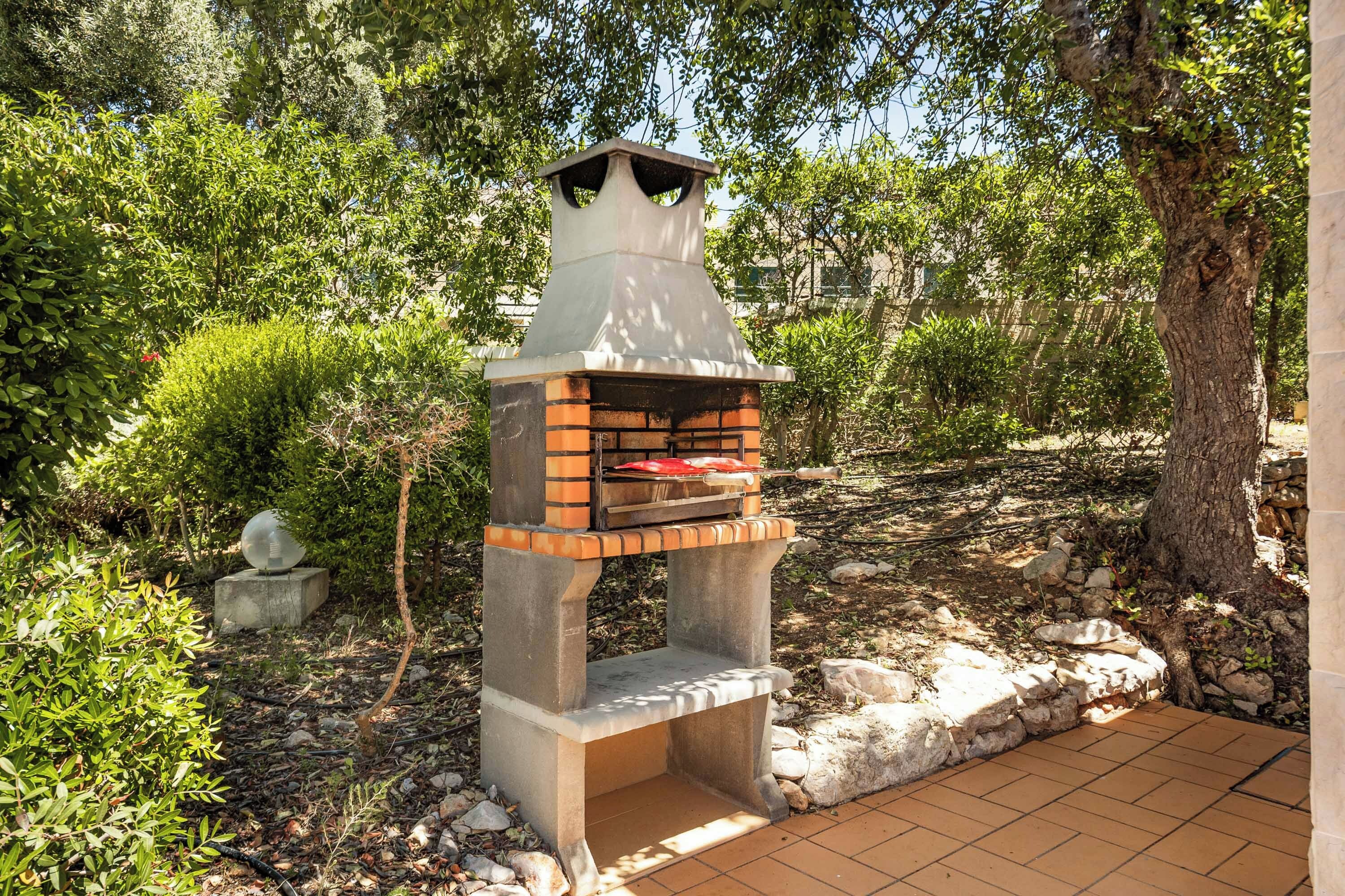 An outdoor brick-built barbecue grill with a chimney, surrounded by greenery and rocks.