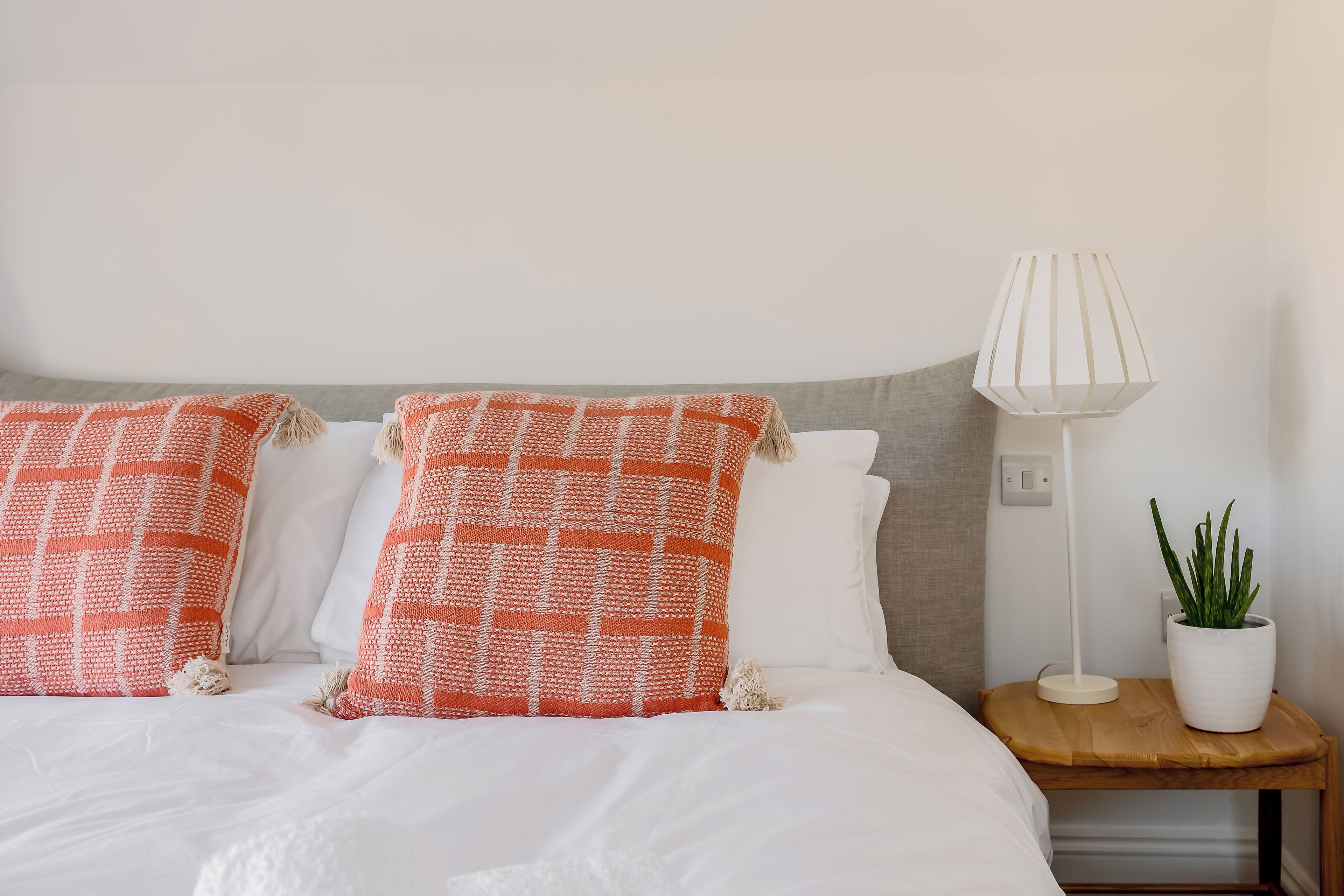 Un lit avec une literie blanche et deux oreillers orange, une lampe blanche posée sur une table de chevet en bois, et une plante en pot.