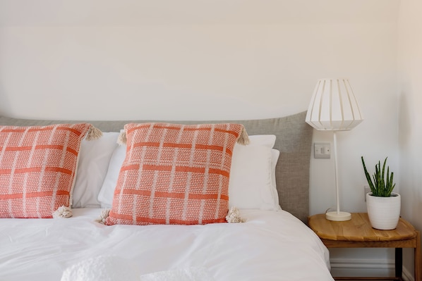 A bed with white bedding and two orange pillows, a white lamp on a wooden nightstand, and a potted plant.