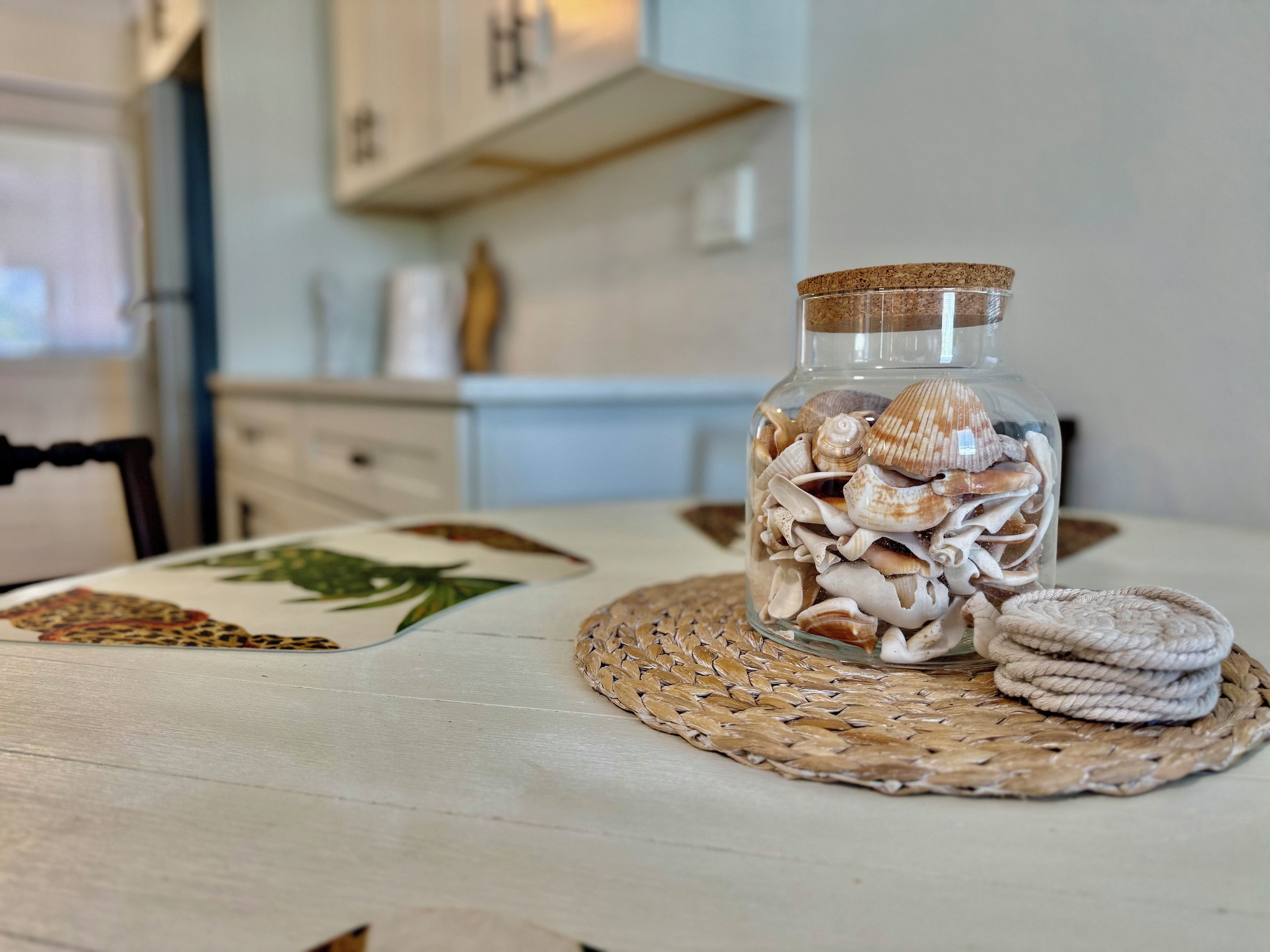 A glass jar filled with seashells on a woven placemat, placed on a table with a patterned placemat.