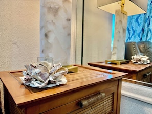 Una mesita de noche de madera con un vaso y conchas decorativas, un espejo que refleja una lámpara y una silla, y una pared con textura al fondo.
