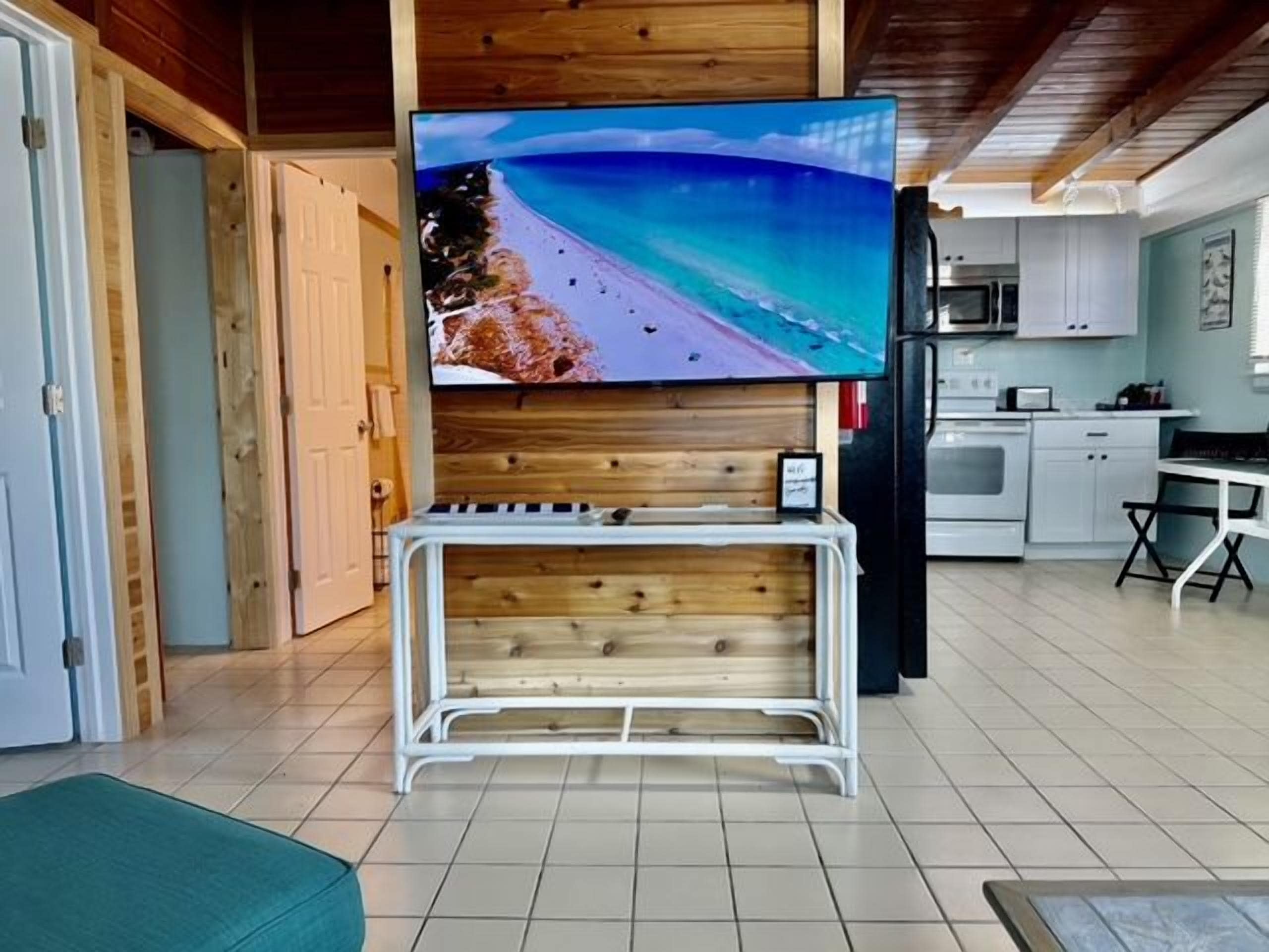 A living room with a television mounted on a wooden wall, a white bench, and a kitchen area in the background.