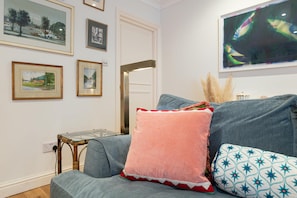 A living room with a grey sofa, a pink cushion, and a patterned pillow. There are framed artworks on the wall and a television screen displaying an image. - Cozy 2Bed Flat with Private Patio, Shepherd’s Bush - Pass the Keys (London)