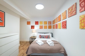 A hotel room with a bed, a bedside table with a lamp, and a wall with multiple artworks. - Cozy 2Bed Flat with Private Patio, Shepherd’s Bush - Pass the Keys (London)