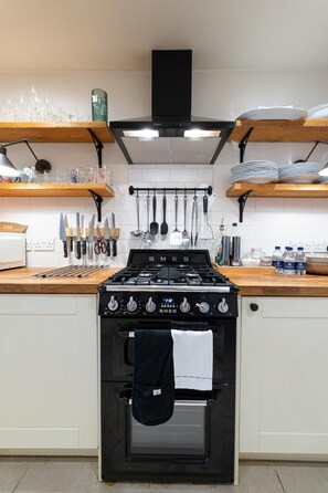 A kitchen with a black stove, white cabinets, and wooden shelves. - Cozy 2Bed Flat with Private Patio, Shepherd’s Bush - Pass the Keys (London)