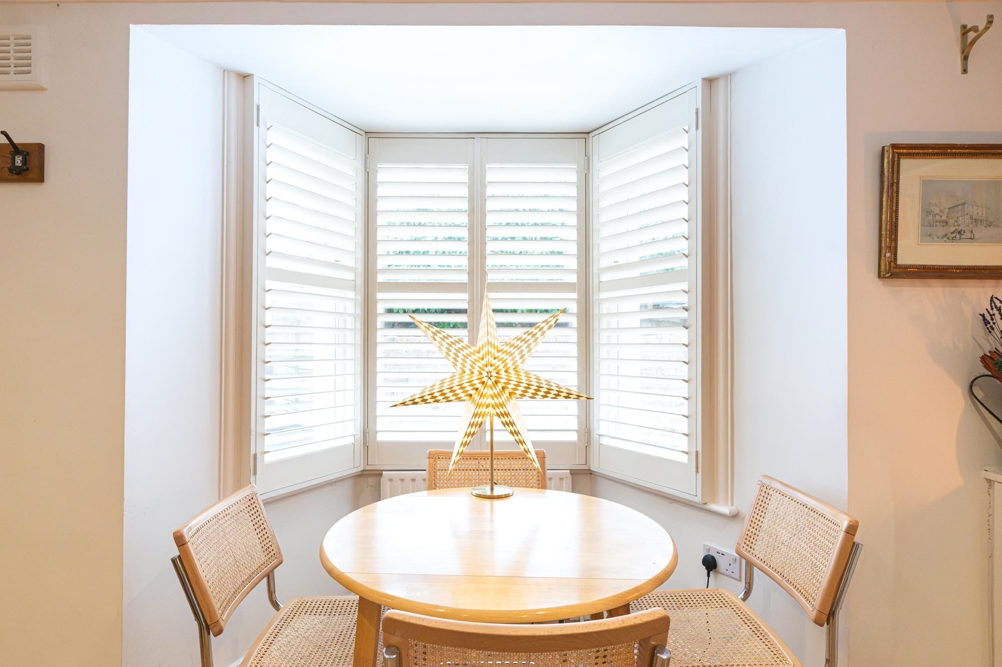 Ein Eckzimmer mit rundem Tisch und vier Korbstühlen, einer sternförmigen Lampe und weißen Fensterläden.