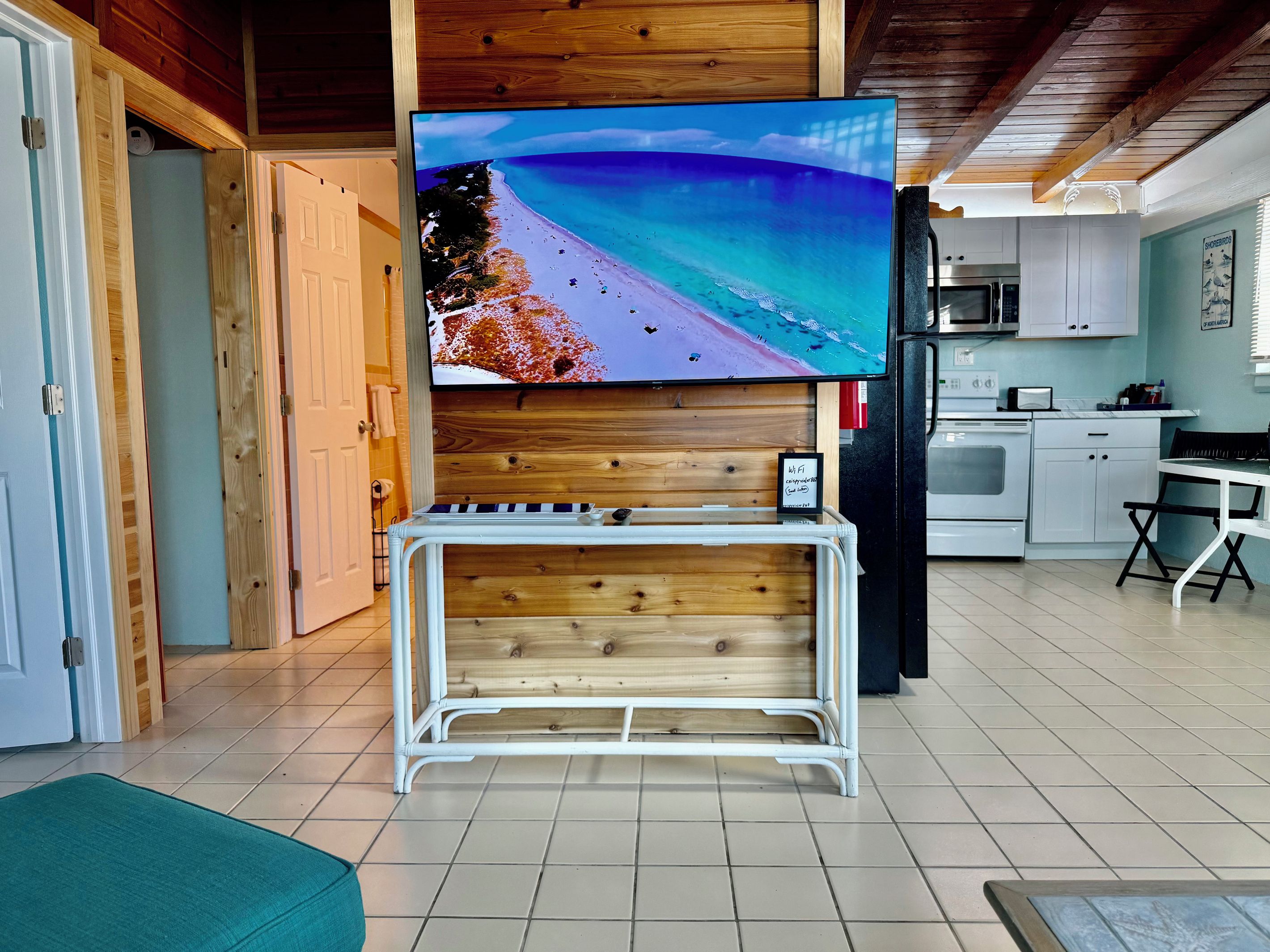 A living room with a flat-screen TV mounted on a wooden wall, a white metal TV stand, and a kitchen area with white cabinets and appliances.