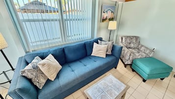 A living room with a blue sofa, patterned armchair, teal ottoman, and a small table with a decorative tile top.