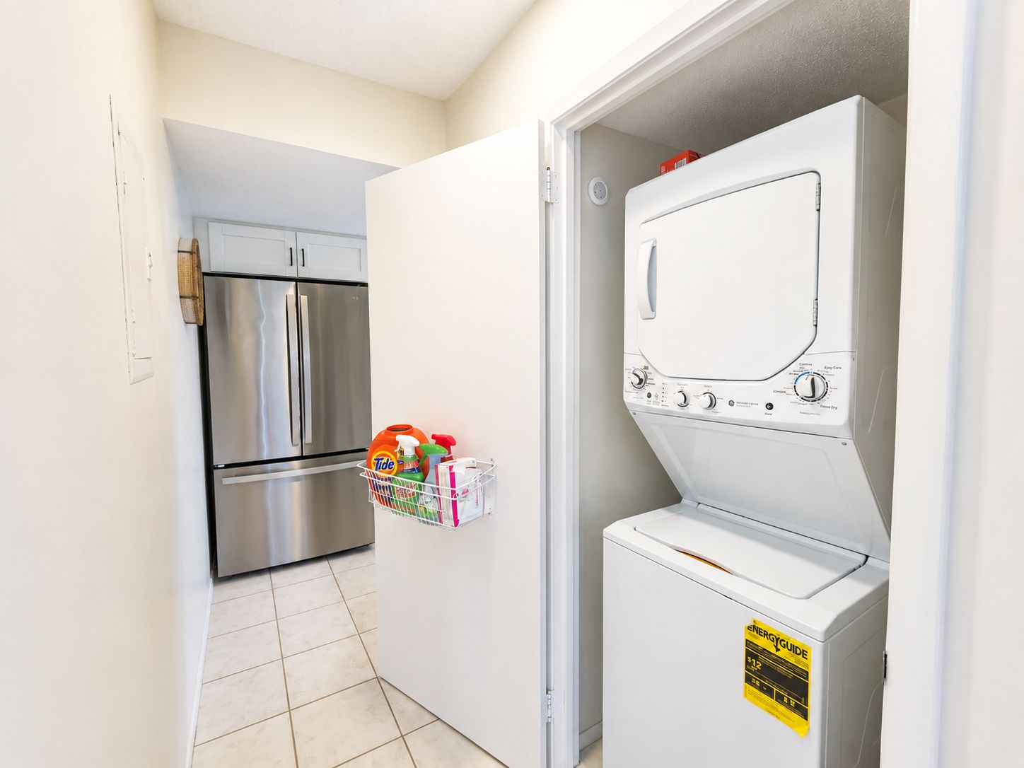 A laundry room with a front-loading washer and dryer, a stainless steel refrigerator, and a basket of laundry supplies on the wall.