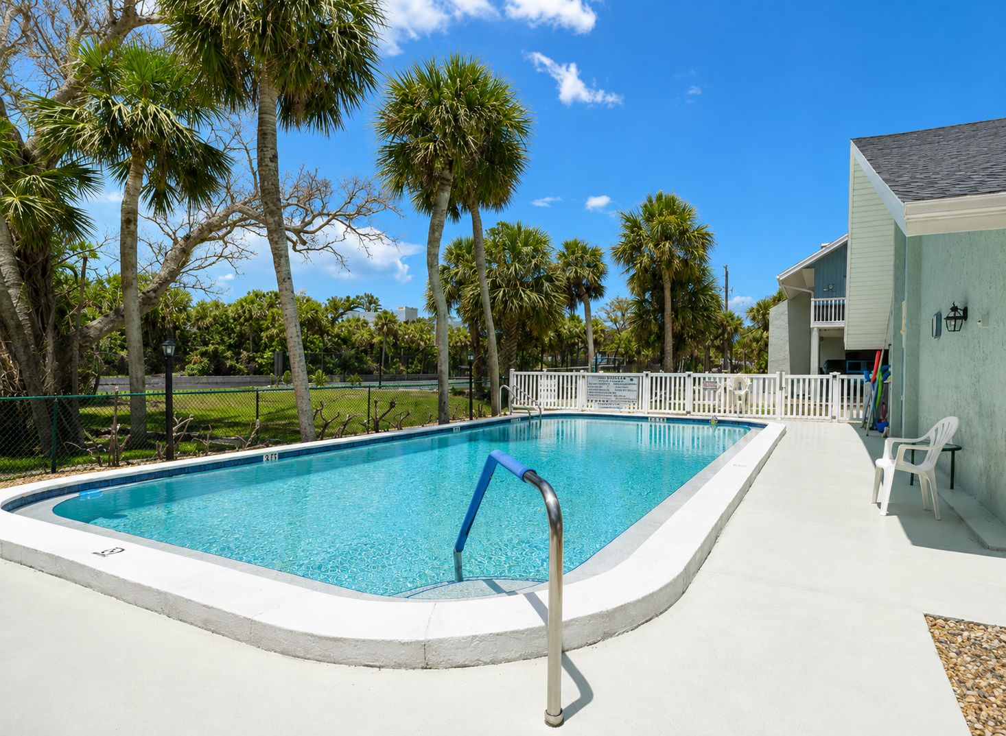 A swimming pool with a white deck, surrounded by palm trees and a fence.