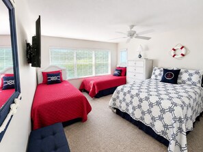 Quarto com duas camas de solteiro, uma cama de casal, ventilador de teto, cômoda e janela com persianas.