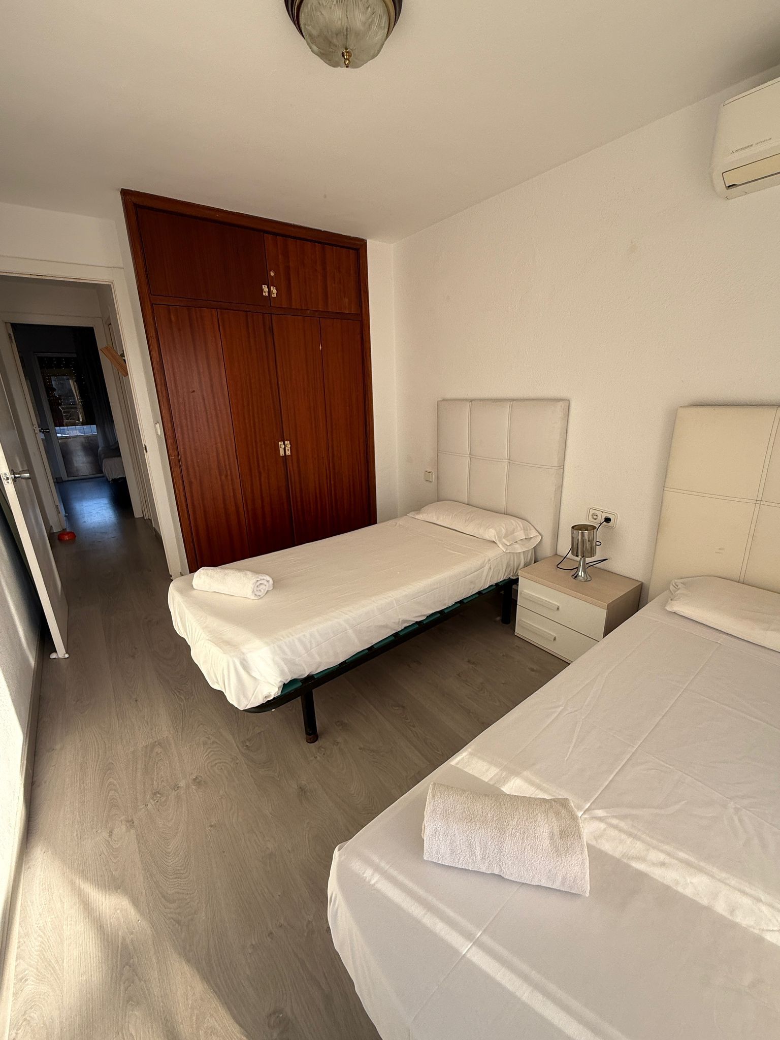 A bedroom with two single beds, a wooden wardrobe, a bedside table with a lamp, and a ceiling light fixture.