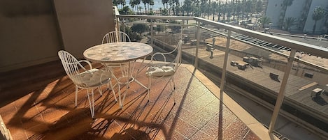 A balcony with a round table and chairs, a view of the beach, and a cityscape in the distance.