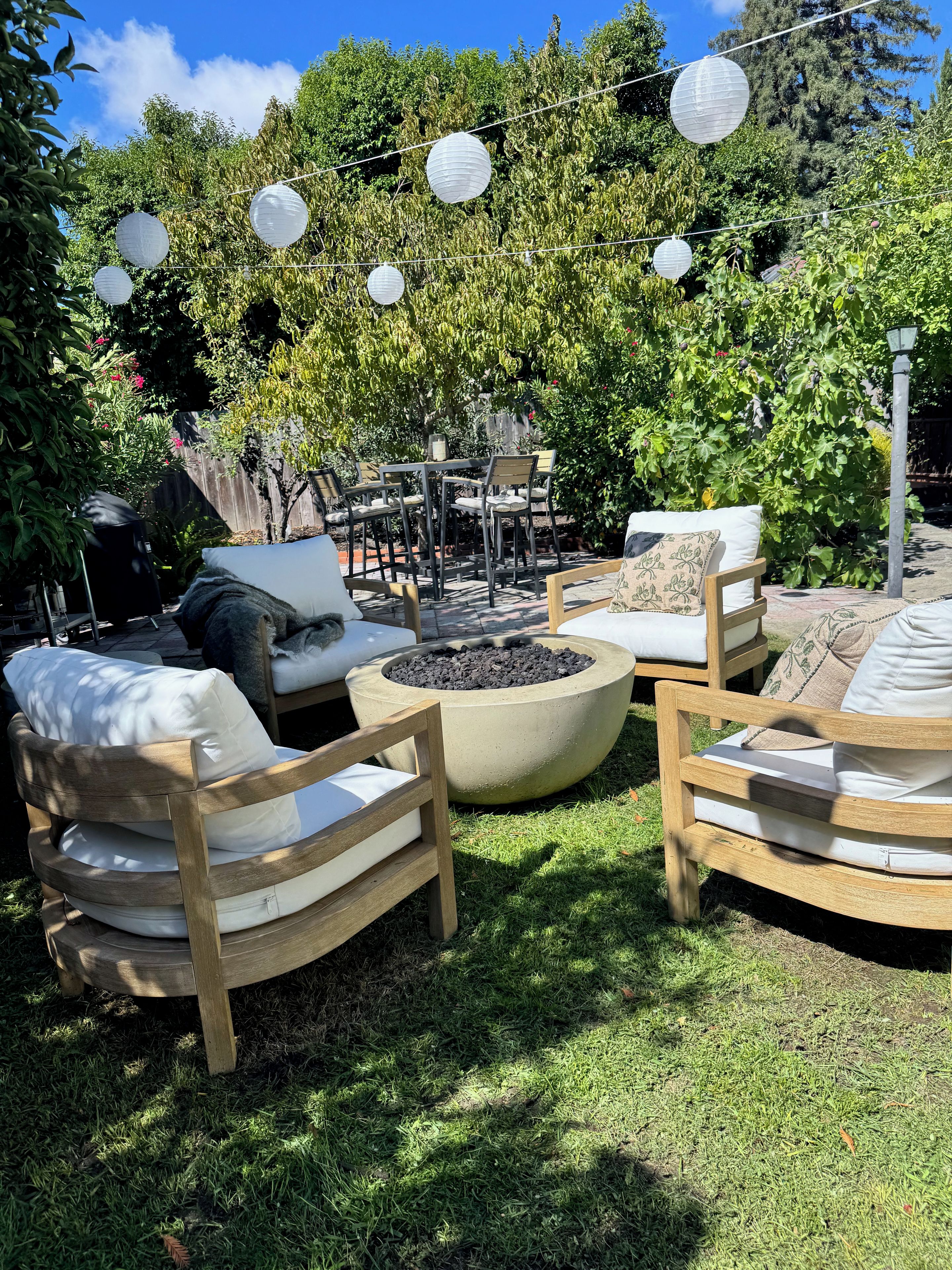 An outdoor seating area with wooden chairs and a round table, surrounded by greenery and string lights.