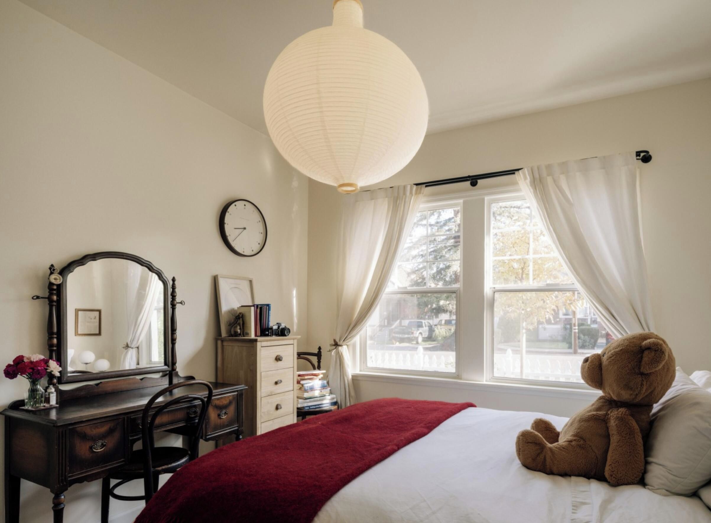 A bedroom with a bed, a dresser, a mirror, a clock, and a window with curtains.