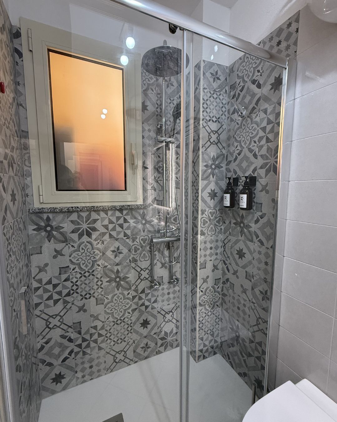 Una ducha con puerta de cristal, un espejo encima y azulejos con diseño en las paredes.