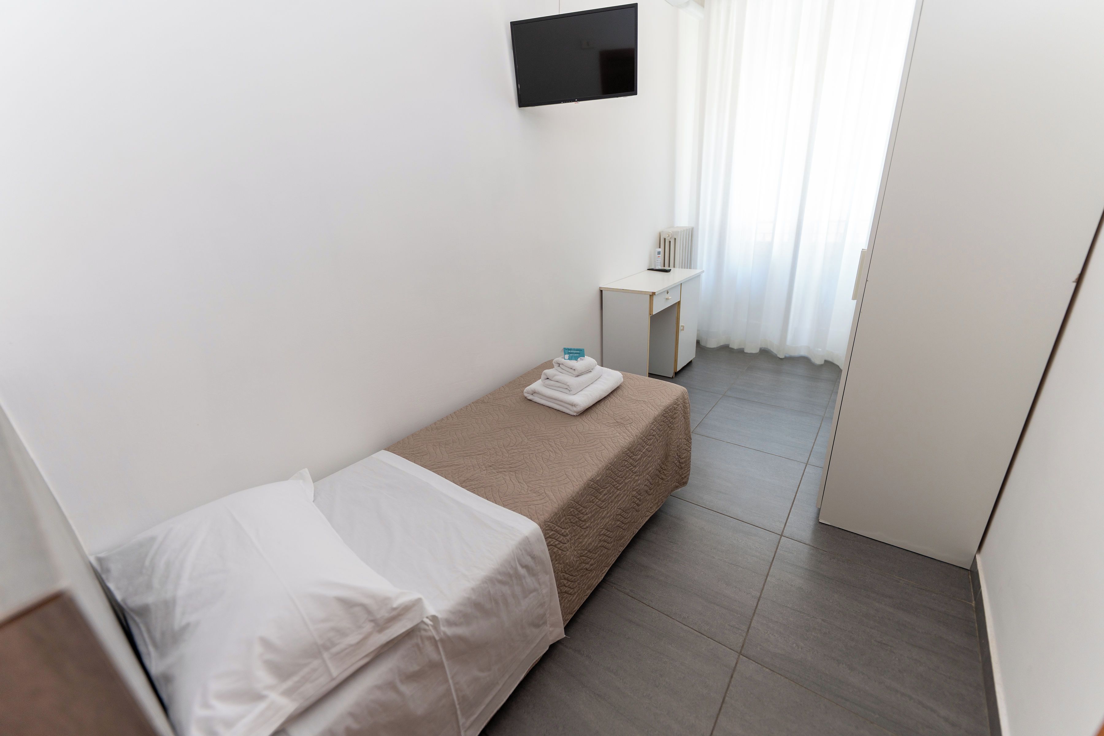 Ein kleiner, sauberer Raum mit einem Einzelbett, einem Nachttisch, einem an der Wand montierten Fernseher und einer Heizung.