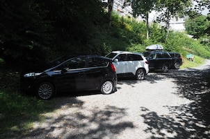 Un camino de grava con coches aparcados, rodeado de vegetación y árboles.