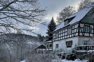 A traditional half-timbered house with a snow-covered roof, surrounded by a forested area.