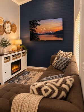 Ein gemütliches Wohnzimmer mit Kamin, einer dunkelblauen Wand, einem bequemen braunen Sofa und einem Bild von einem Sonnenuntergang in einem Rahmen.