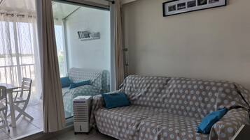 A room with a sofa, a table, and a chair. There is a window with curtains, a wall-mounted shelf with books, and a framed picture.
