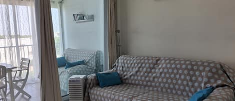 Une pièce comprenant un canapé, une table et une chaise. Il y a une fenêtre avec des rideaux, une étagère fixée au mur avec des livres et un tableau encadré.