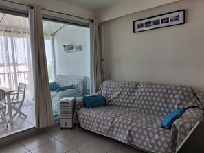 A room with a sofa, a table, and a chair. There is a window with curtains, a wall-mounted shelf with books, and a framed picture. - Seafront Stay Near South Beach (Le Grau-Du-Roi)