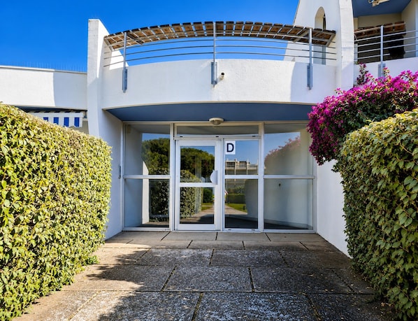 Entrada de um prédio com porta de vidro identificada pela letra 'D', cercada por vegetação e céu azul límpido.