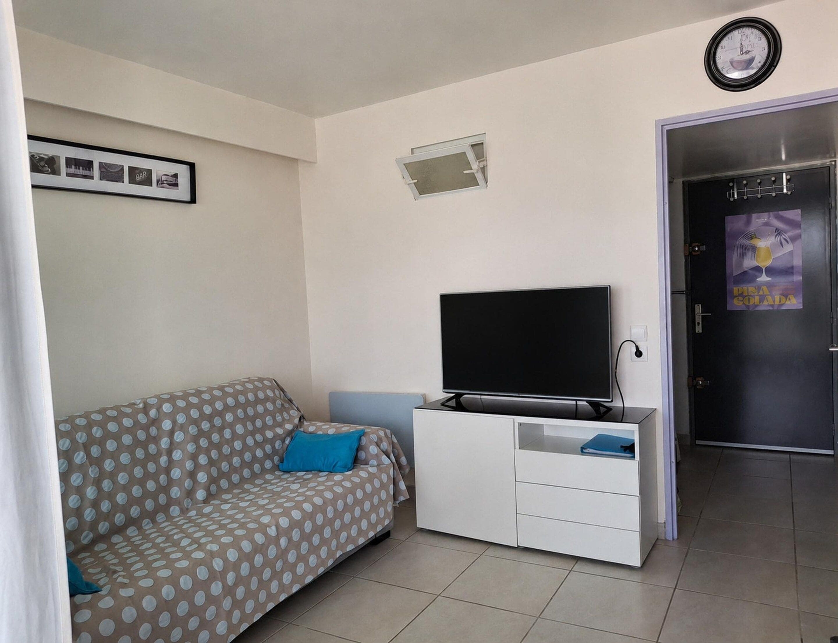 A living room with a sofa, a flat-screen TV, a white cabinet, a clock, and a door with a poster.