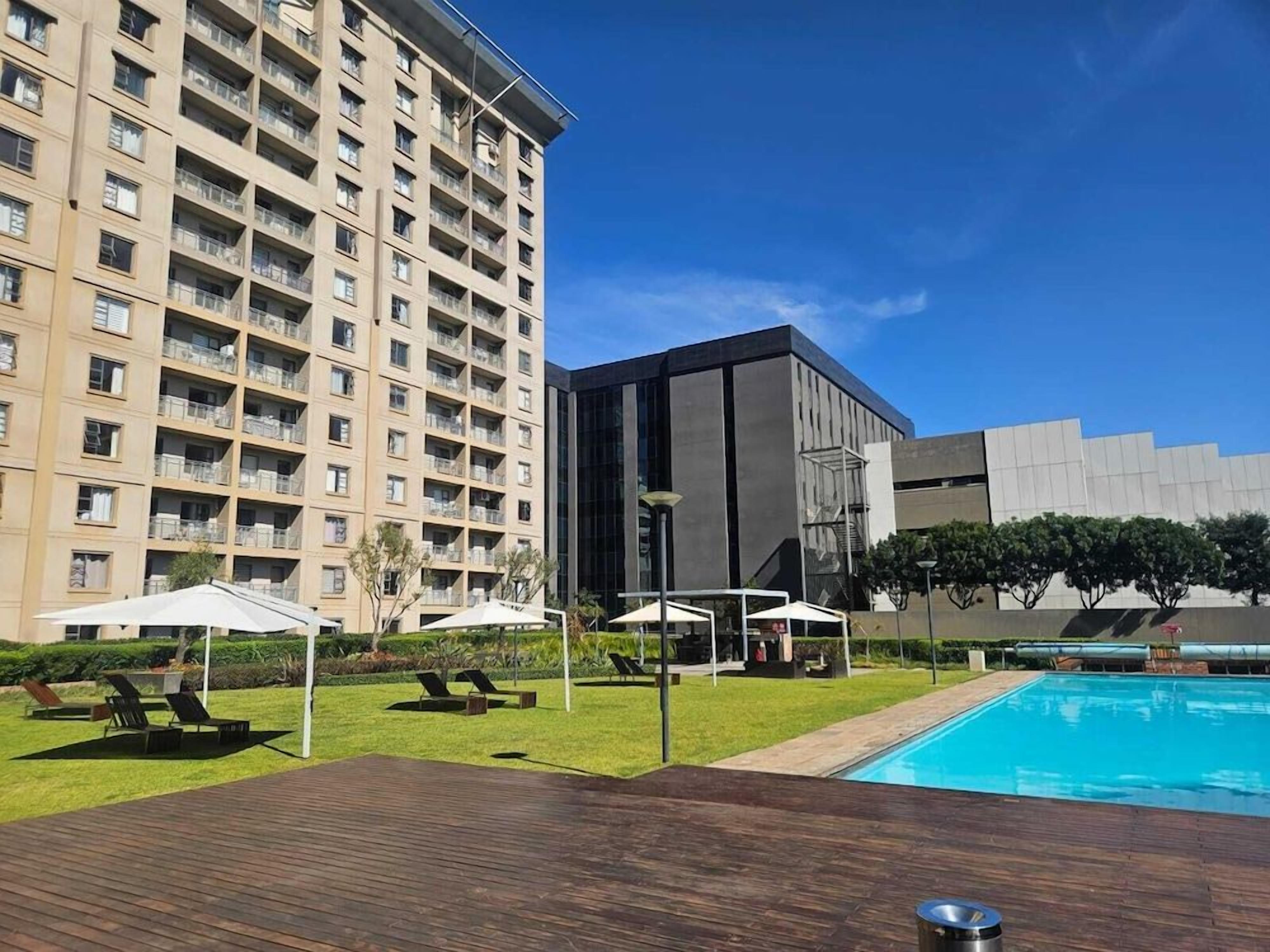 Un espace piscine d’hôtel avec une terrasse en bois, des chaises longues, des parasols et un grand bâtiment en arrière-plan.
