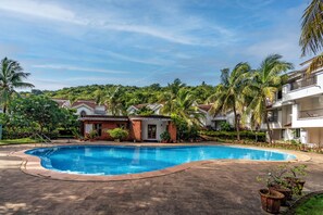 A swimming pool surrounded by buildings with balconies, palm trees, and a hilly backdrop.