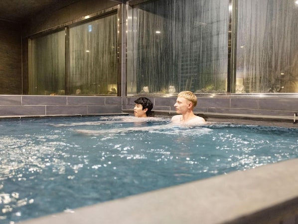A swimming pool with two individuals in the water, surrounded by a tiled wall and large windows.
