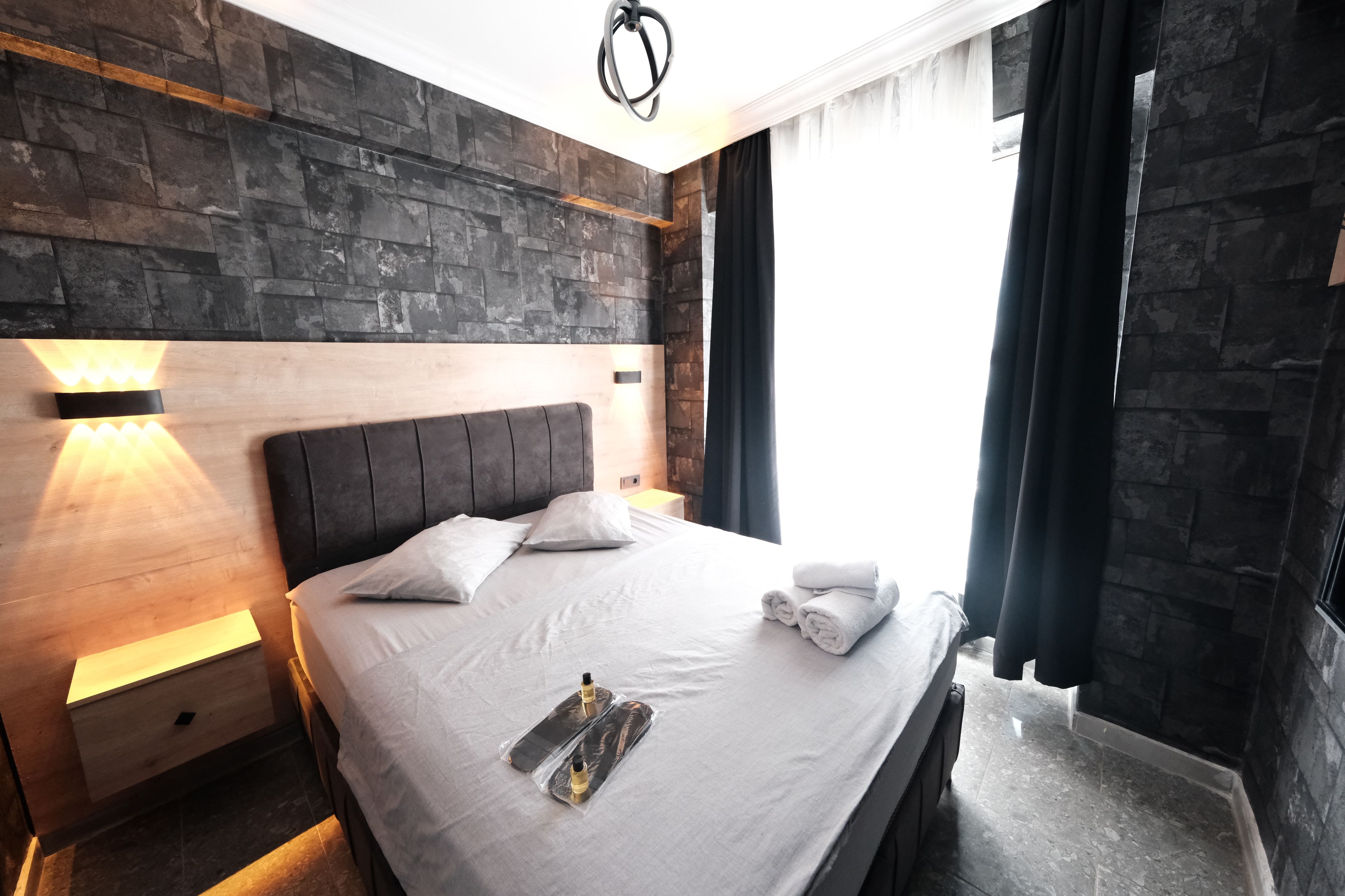 A modern hotel room with a stone wall, a wooden headboard, a neatly made bed with white linens, a bedside lamp, and a window with curtains.