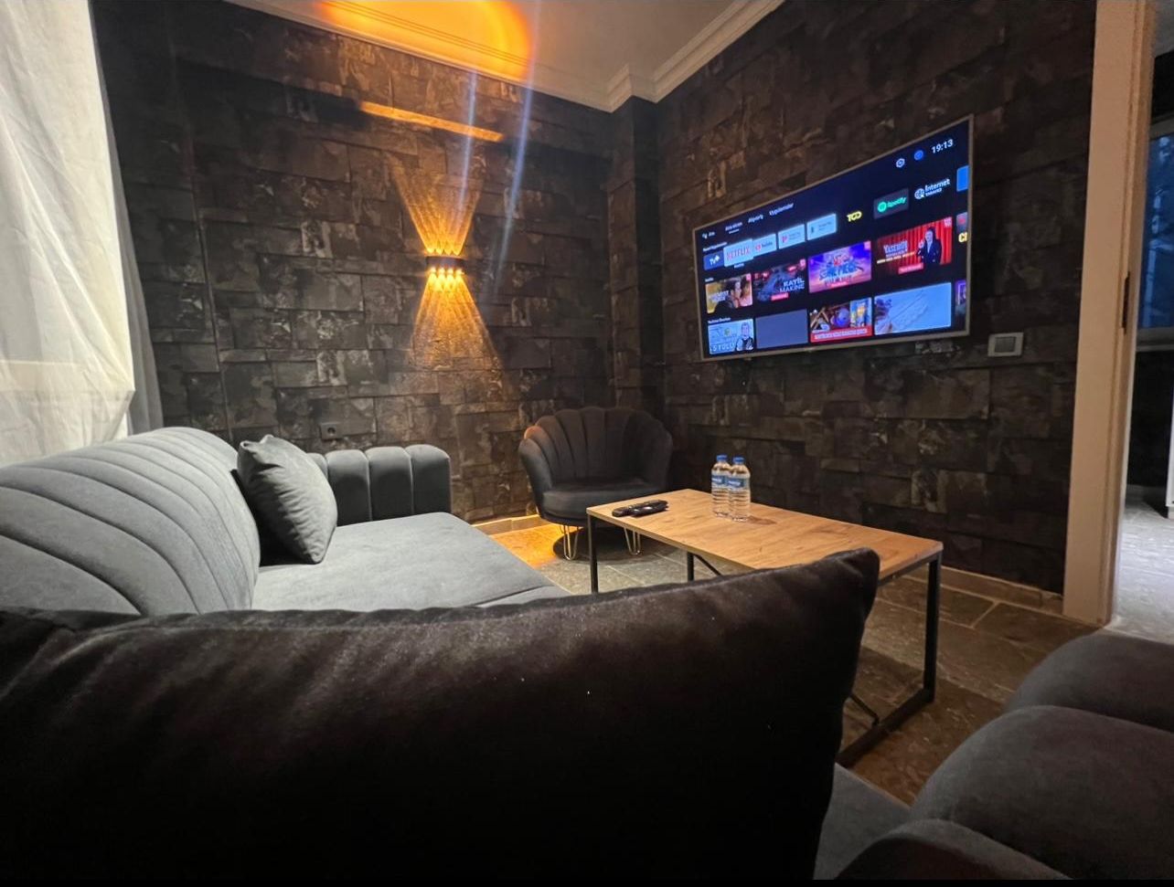 Una sala de estar moderna con una pared de piedra, un televisor de pantalla plana montado en la pared, un sofá gris, una mesa de centro de madera y un sillón negro.