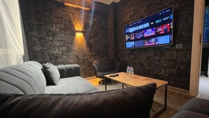 A modern living room with a stone wall, a flat-screen TV mounted on the wall, a grey sofa, a wooden coffee table, and a black armchair. - Luna Suites Hotel (Eskişehir)