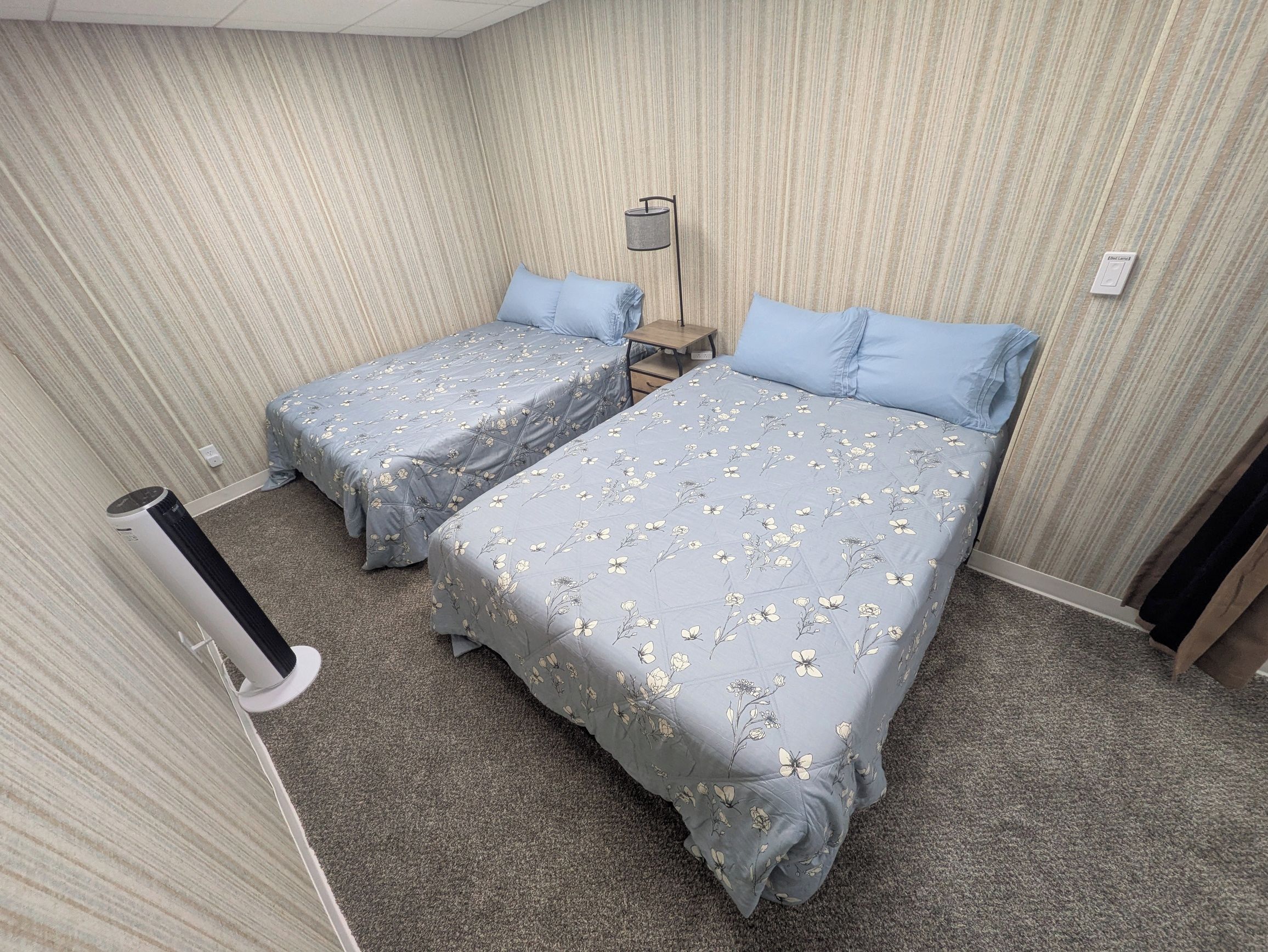 A hotel room with two beds, a bedside table, a lamp, and a wall-mounted light switch.