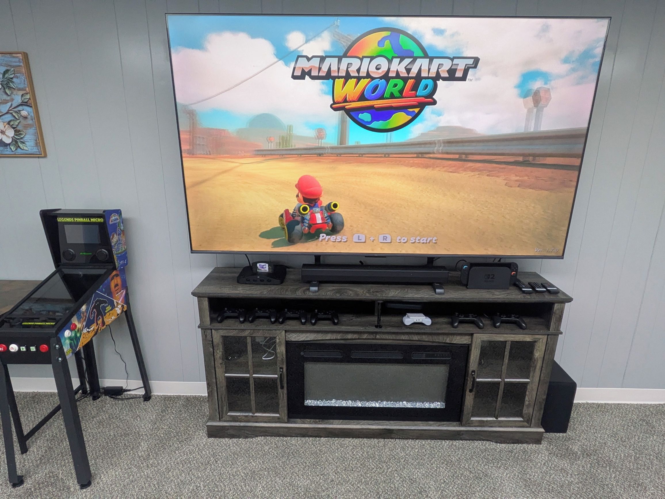Een televisie waarop Mario Kart World te zien is, met een spelconsole en controllers op een houten dressoir.