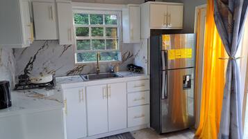Cocina compacta con armarios blancos, refrigerador de acero inoxidable, ventana con vistas a vegetación y una puerta con cortinas.