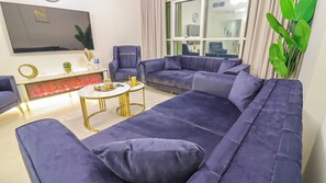 Een moderne woonkamer met een grote bank, een kleinere bank, een salontafel en een fauteuil. Er is een open haard met glazen deuren, een aan de muur bevestigde televisie en een groot raam met gordijnen.