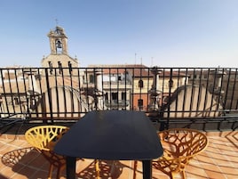 A terrace with a table and chairs, a metal fence, and a view of buildings including a church tower.
