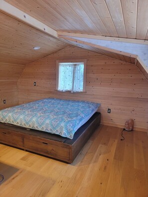 A small, single-room attic bedroom with a wooden sloped ceiling, a window, a bed with a patterned bedspread, and a wooden floor. - Beautiful cabin located close to everything with (EU) King size mattress in upstairs loft, large jetted tub and views of Grewing Glacier and Kachemak Bay from the windows. (Homer)