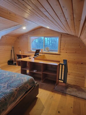 A cozy attic bedroom with a wooden desk, a bed with a patterned bedspread, and a window with a view of trees.